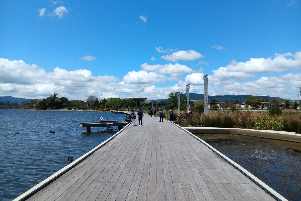 Una larga pasarela de madera se adentra junto al agua del lago Rotorua bajo un cielo azul con nubes dispersas